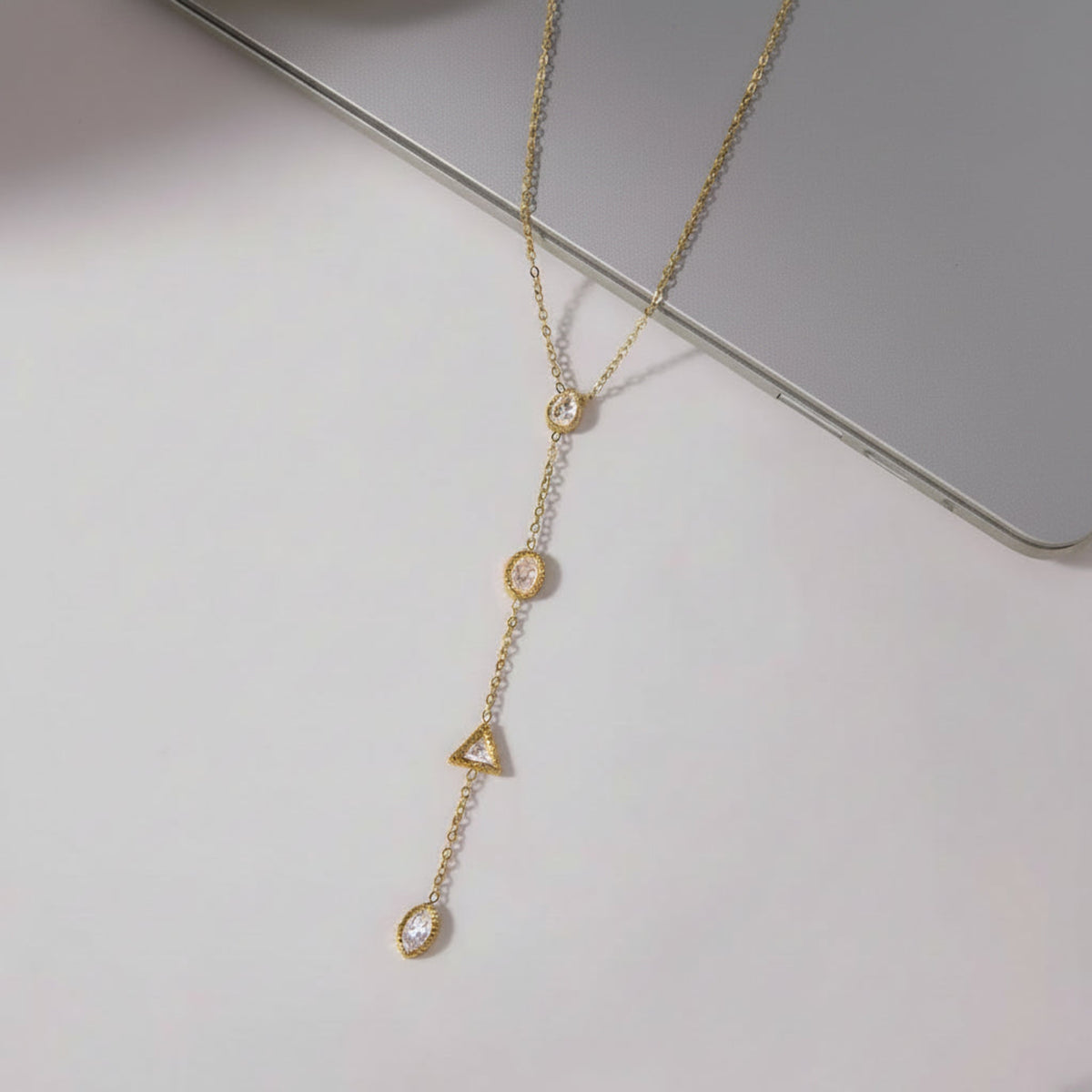 A gold-toned lariat-style necklace with four dangling geometric charms, including a teardrop, circle, triangle, and oval, each accented with small clear stones. The necklace is displayed on a light gray surface alongside a silver laptop and cosmetic tubes.