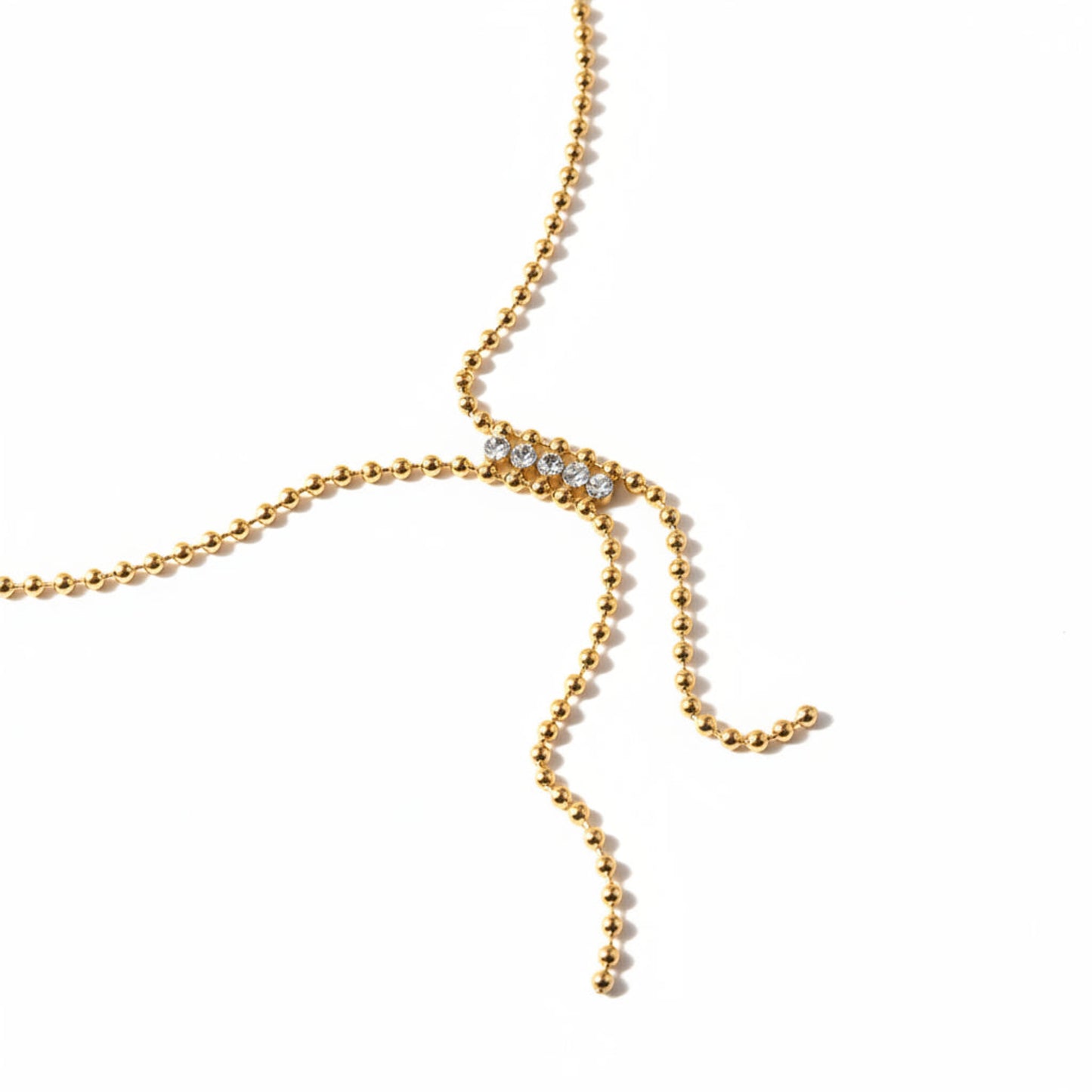 A delicate gold beaded chain necklace with a central row of clear rhinestones, laid out on a warm beige background.