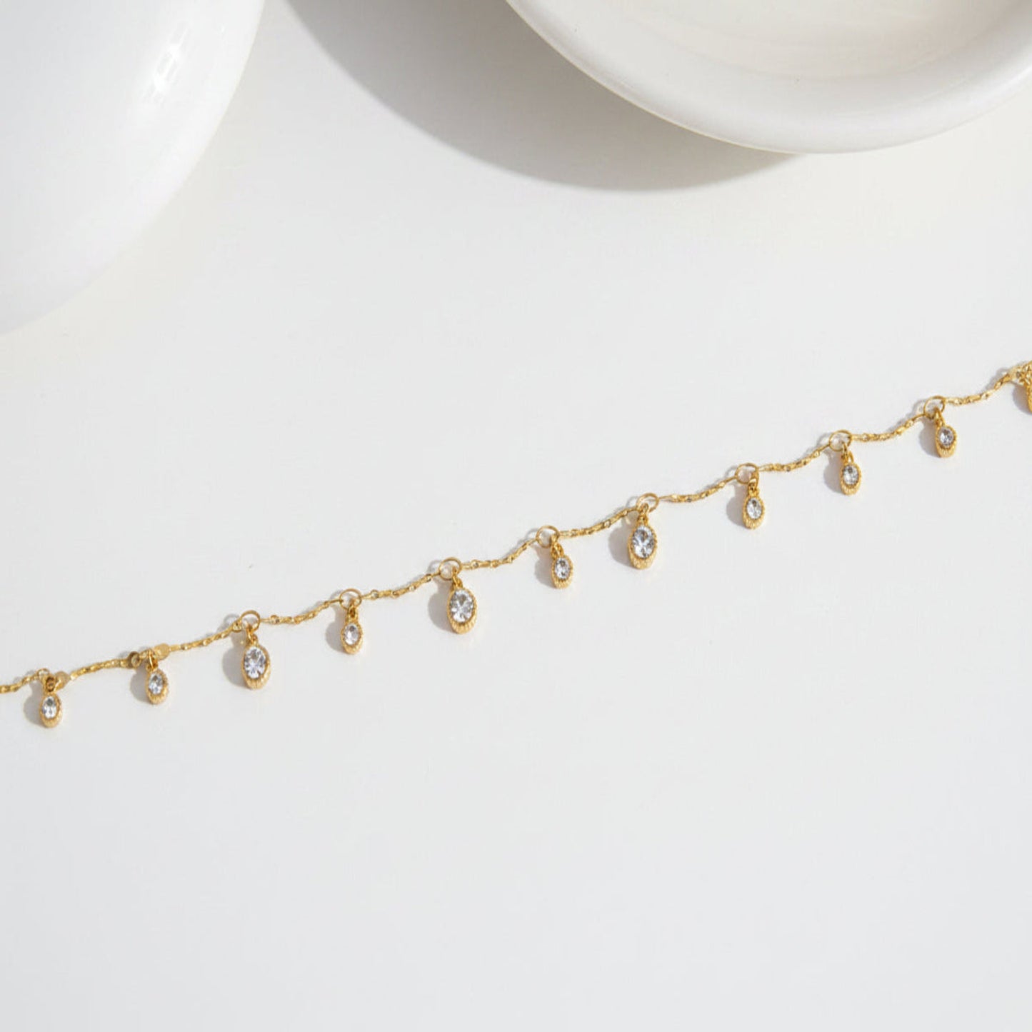 A delicate gold chain bracelet with small teardrop-shaped clear crystal charms, laid out diagonally on a clean white surface.