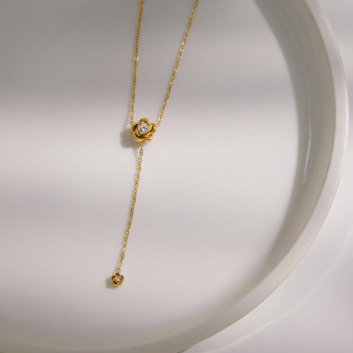 A delicate gold Y-necklace with a rose-shaped pendant and a small drop pendant, both featuring clear gemstones, displayed on a white ceramic dish.