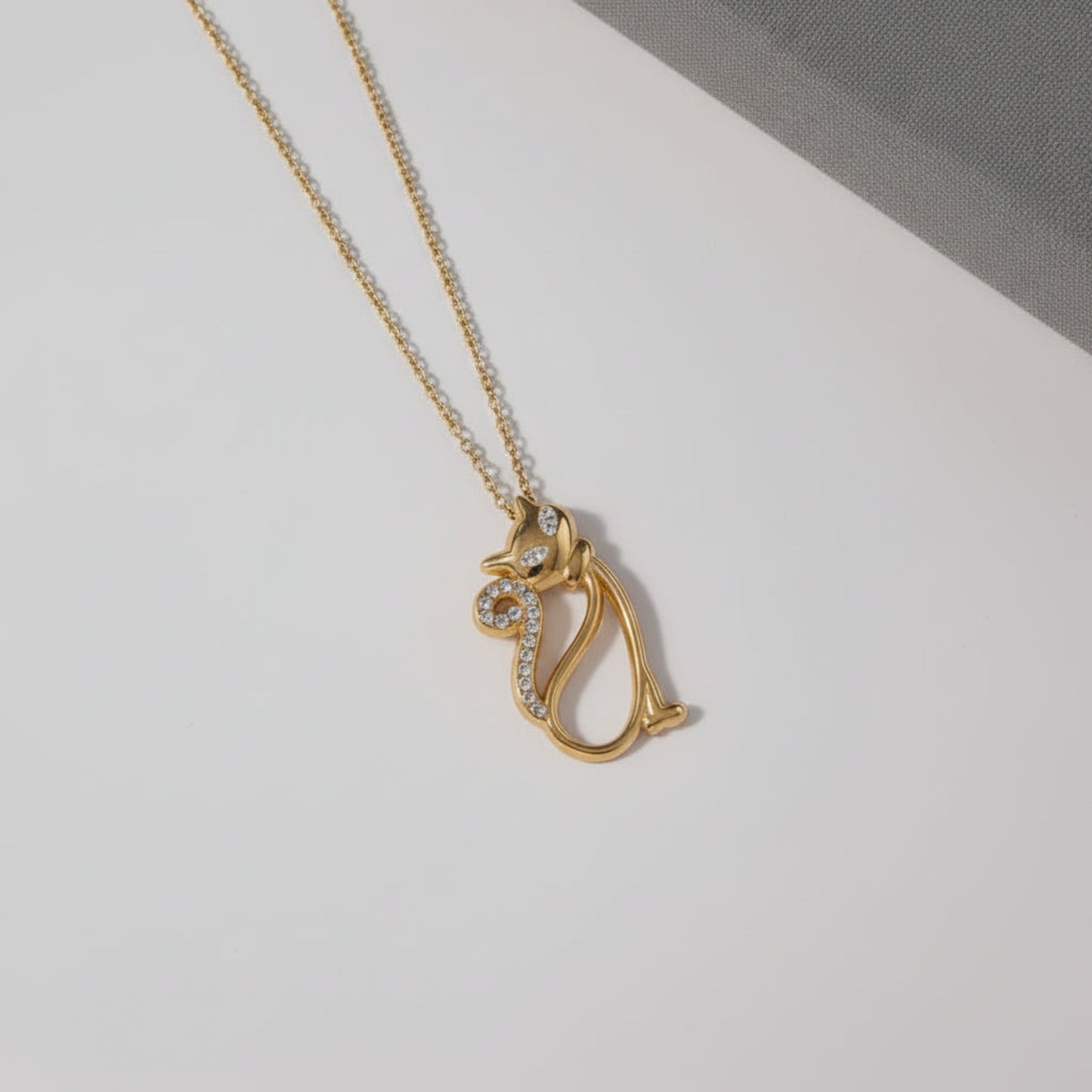 A gold pendant necklace featuring a stylized cat silhouette adorned with small clear rhinestones, displayed on a white surface with a gray textured fabric in the corner.