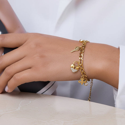 A gold charm bracelet with a key, heart, and flower design, worn on a person's wrist. The bracelet features a gold chain with small charms, including a key, a heart with tiny clear stones, and a flower, set against a white shirt background.