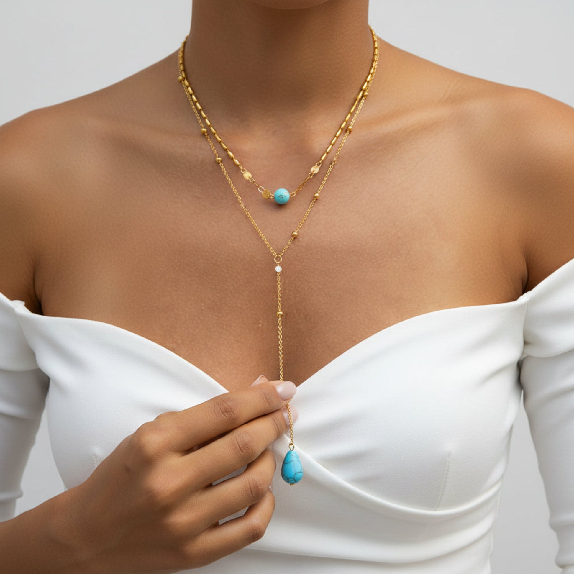 A close-up of a model wearing a layered gold necklace with turquoise blue bead and teardrop pendant, paired with a white off-the-shoulder top.