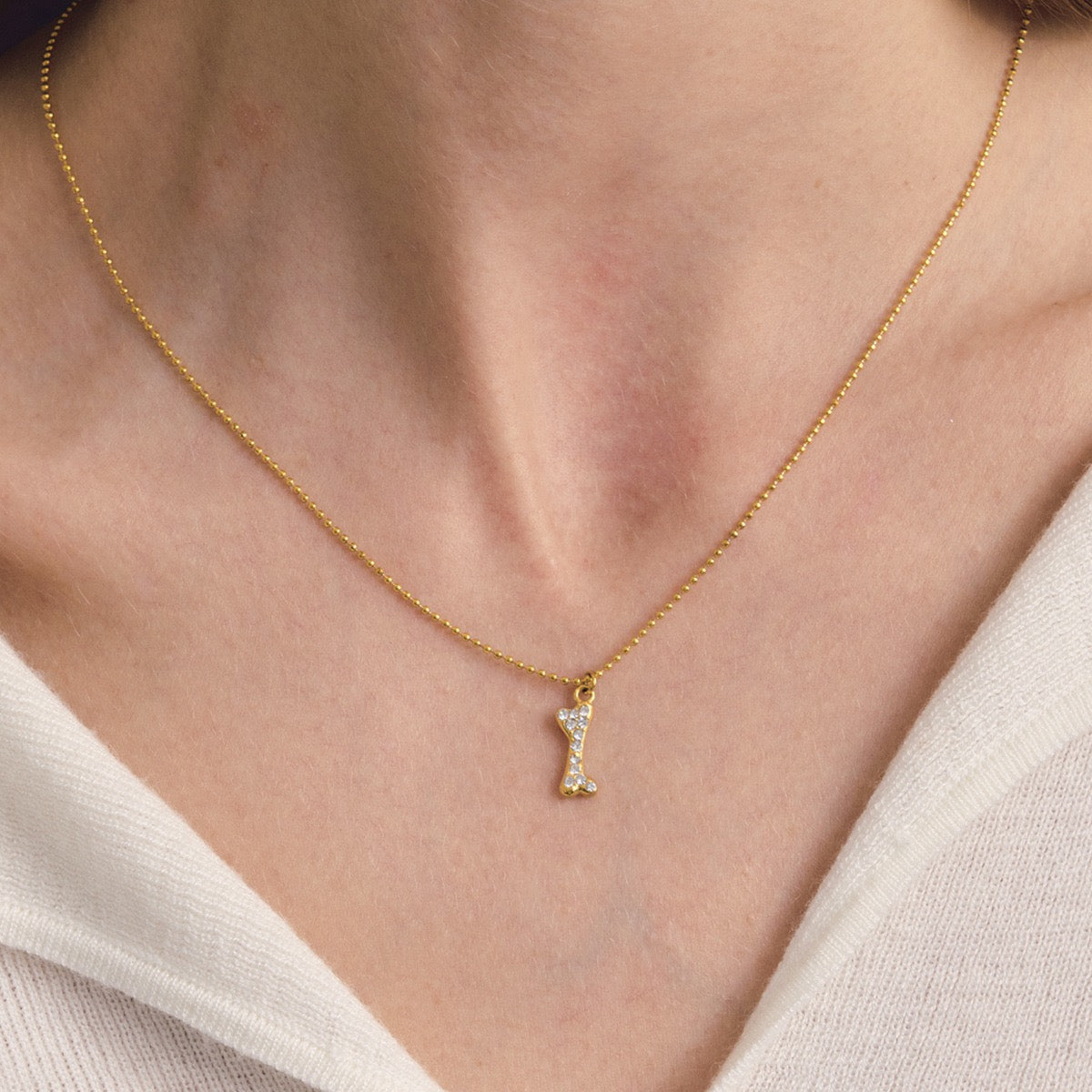 A delicate gold chain necklace with a small bone-shaped pendant adorned with tiny clear stones, worn on a model's neck over a white V-neck top.