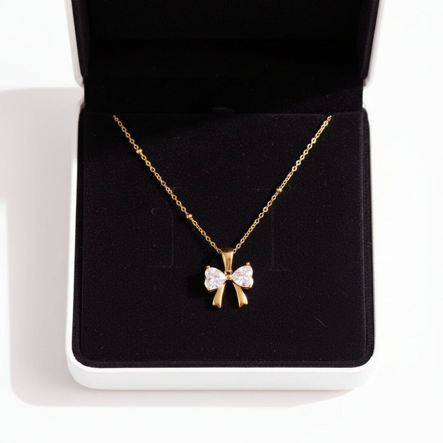 A gold necklace with a bow-shaped pendant set with clear gemstones, displayed inside an open black jewelry box on a white surface.