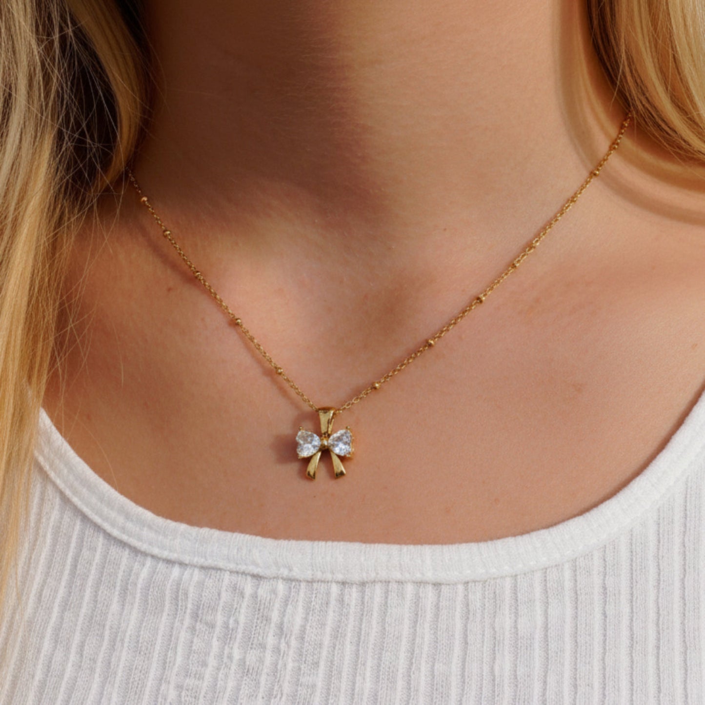 A gold chain necklace with a small bow-shaped pendant set with clear rhinestones, worn on a model's neck over a white ribbed top.