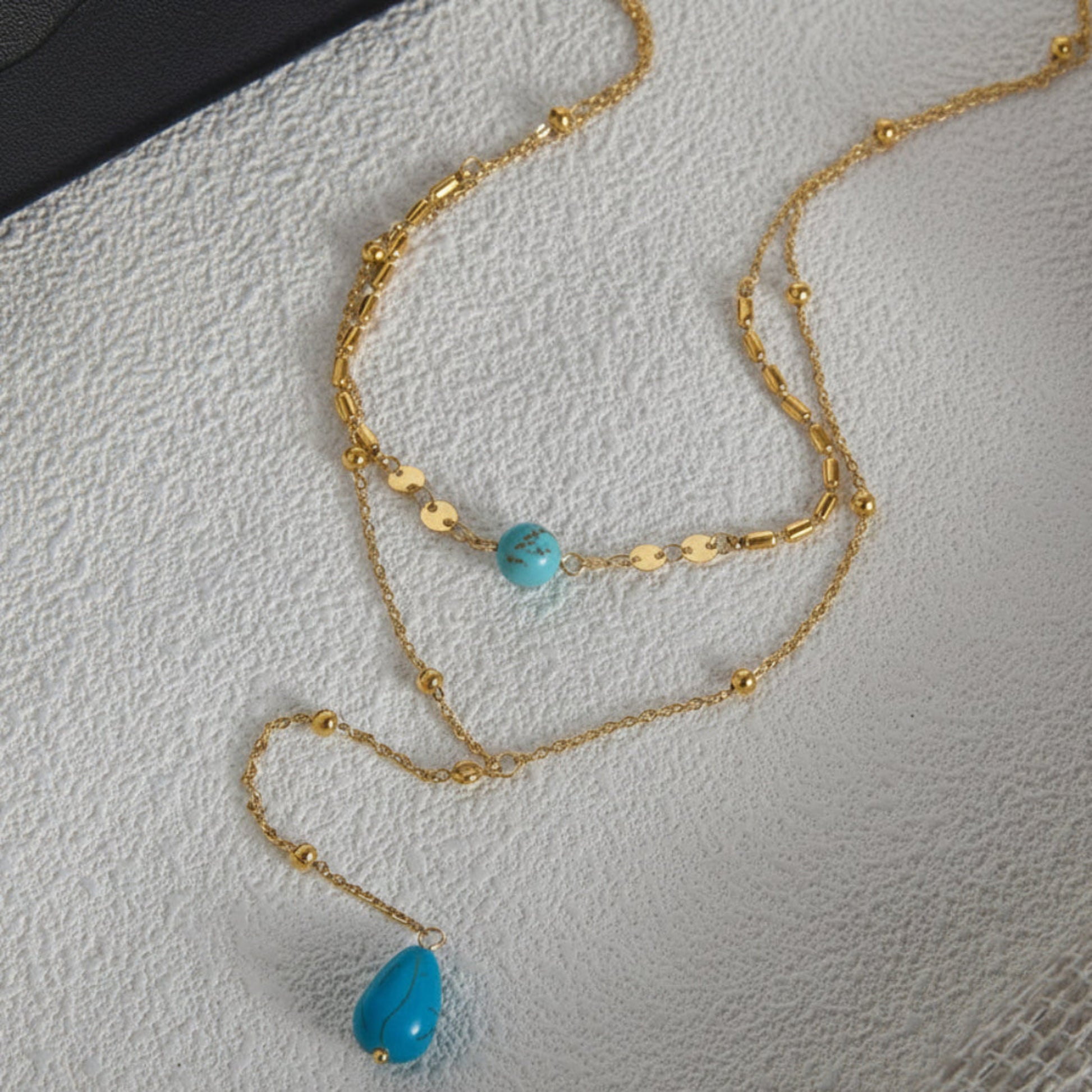 A gold chain necklace with a small turquoise bead and a larger teardrop-shaped turquoise pendant, displayed on a textured light gray surface.