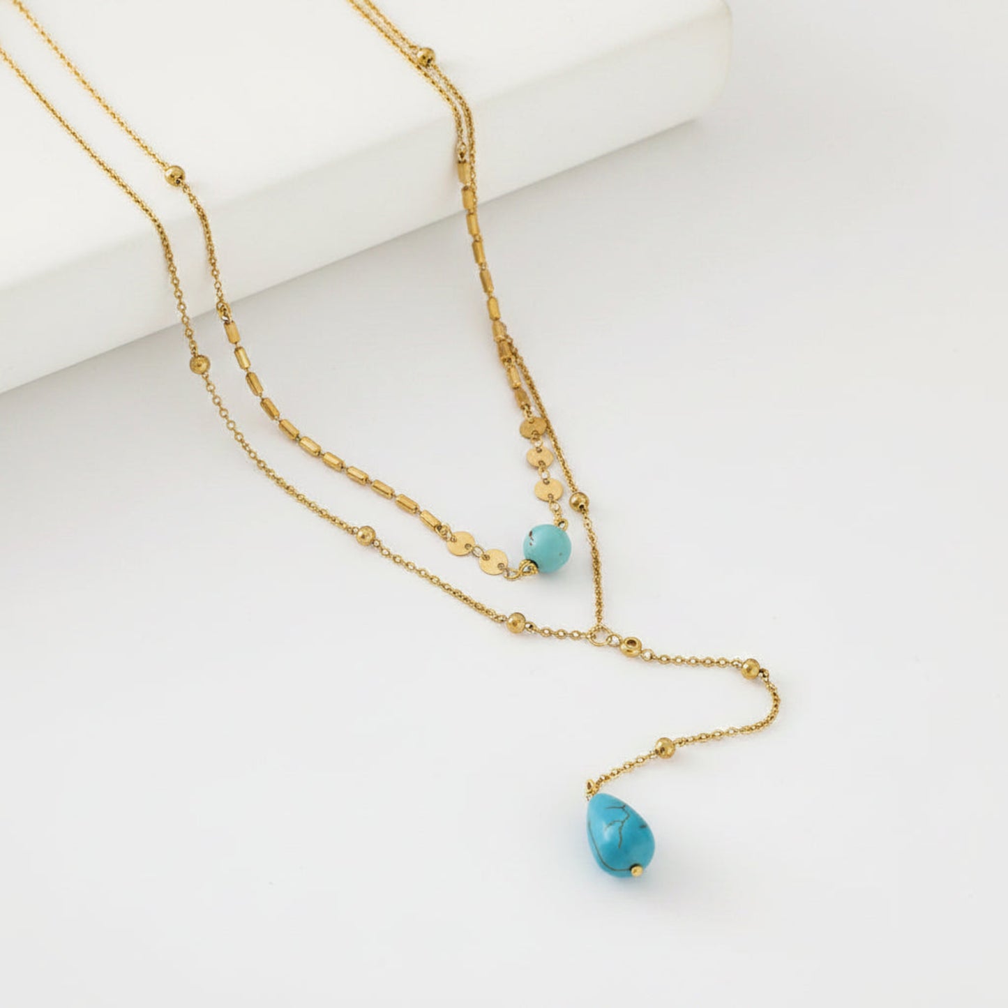 A gold layered necklace with two strands, featuring small gold beads and two turquoise blue teardrop-shaped beads, displayed on a clean white background.