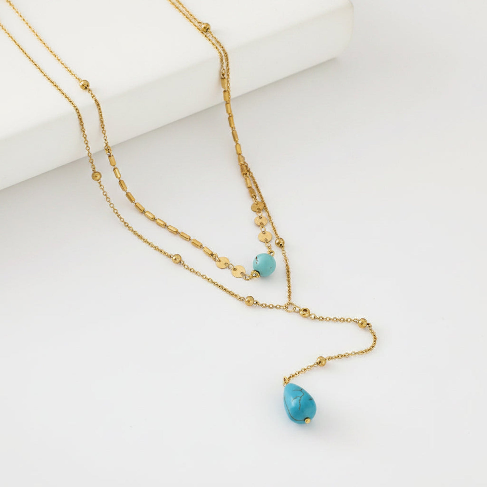 A gold layered necklace with two strands, featuring small gold beads and two turquoise blue teardrop-shaped beads, displayed on a clean white background.