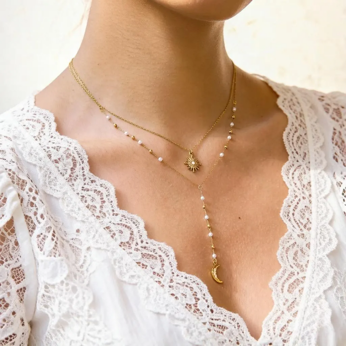 A close-up of a model wearing a layered gold necklace set with pearl accents, a sunburst pendant, and a crescent moon charm, paired with a white lace top.