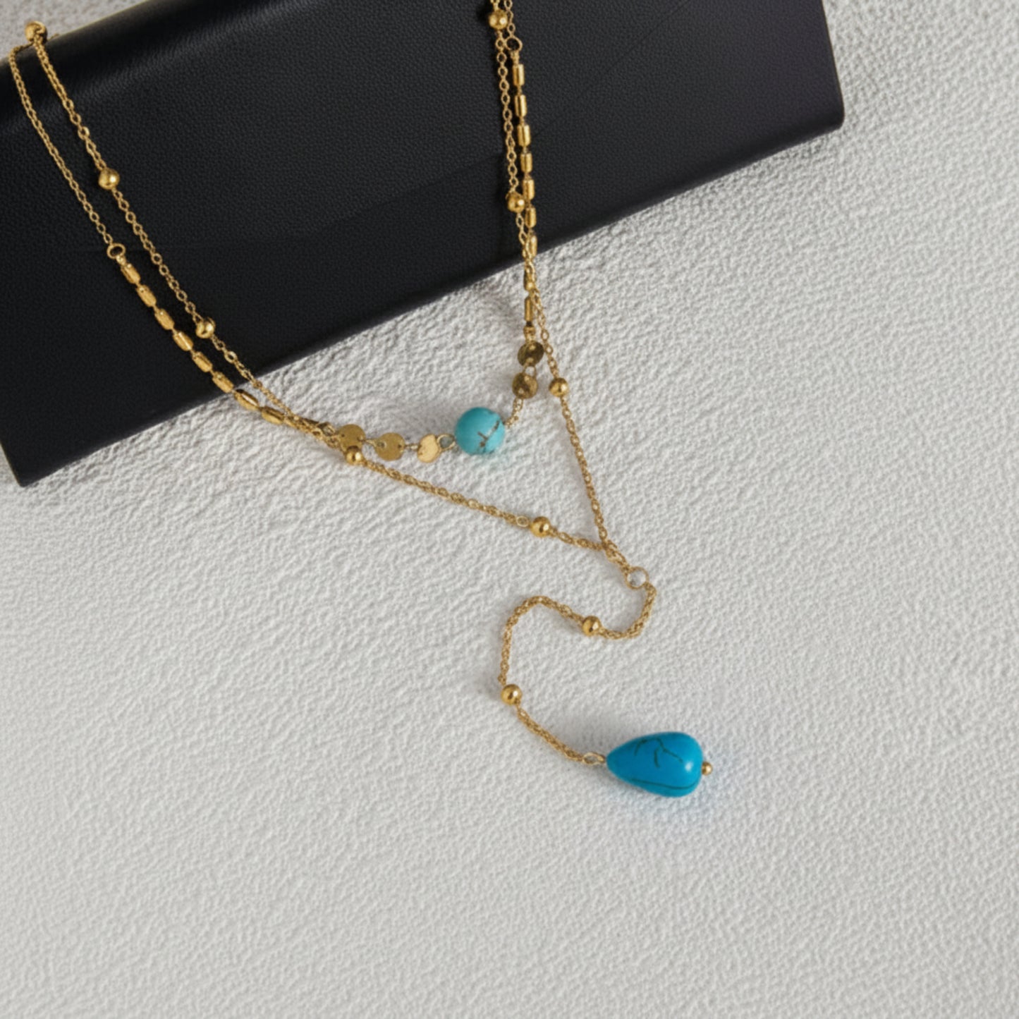 A gold-tone double-strand necklace with turquoise beads and a teardrop-shaped turquoise pendant, displayed on a textured white surface next to a black  jewelry box.