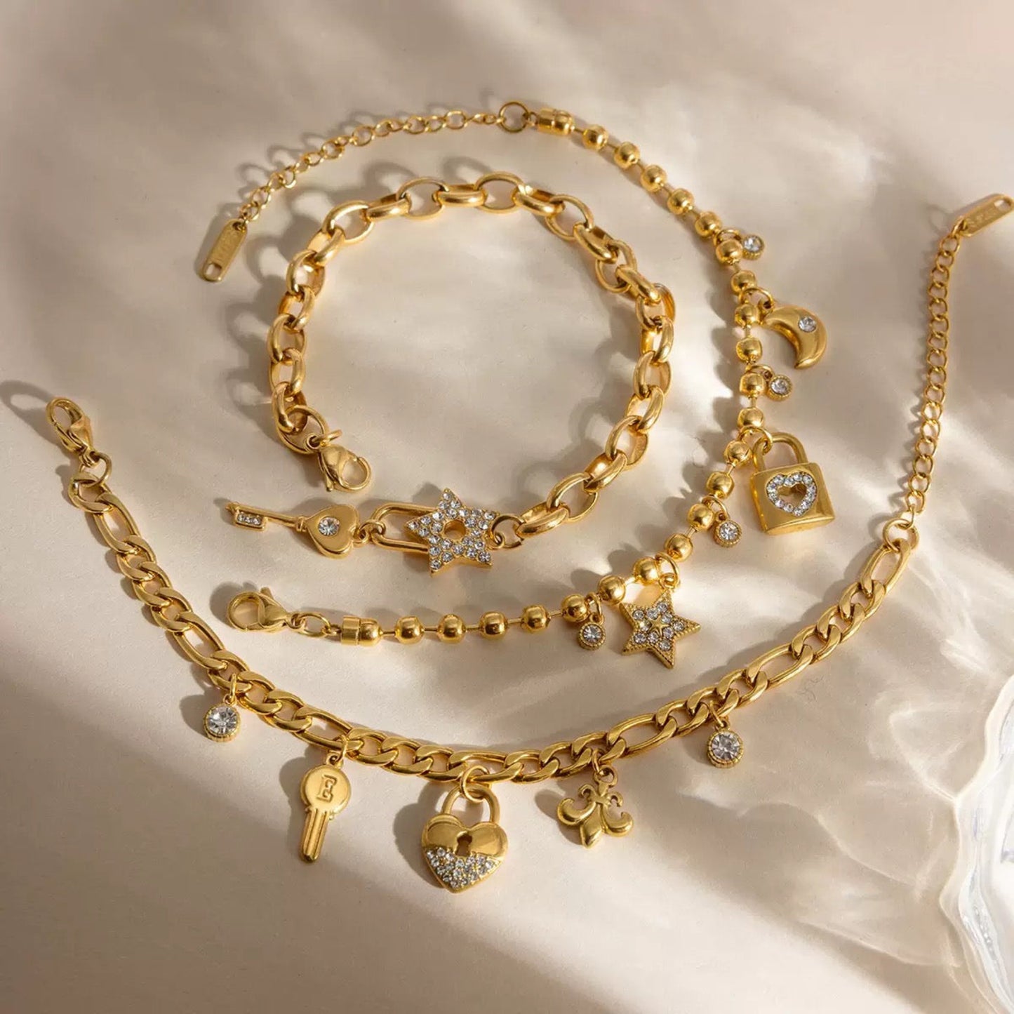 A set of gold-toned chain bracelets with charms, including stars, hearts, a padlock, a key, and moon motifs, arranged on a soft beige fabric background.