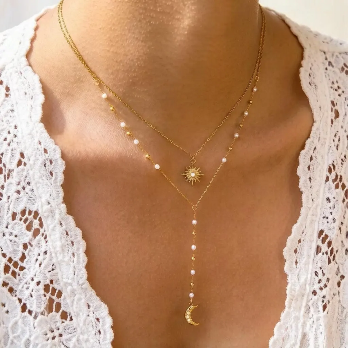 A close-up of a person's neck and chest wearing a layered gold necklace set. The necklace features a sunburst pendant, a long drop chain with small white beads, and a crescent moon pendant, layered over a delicate chain with small white pearls.